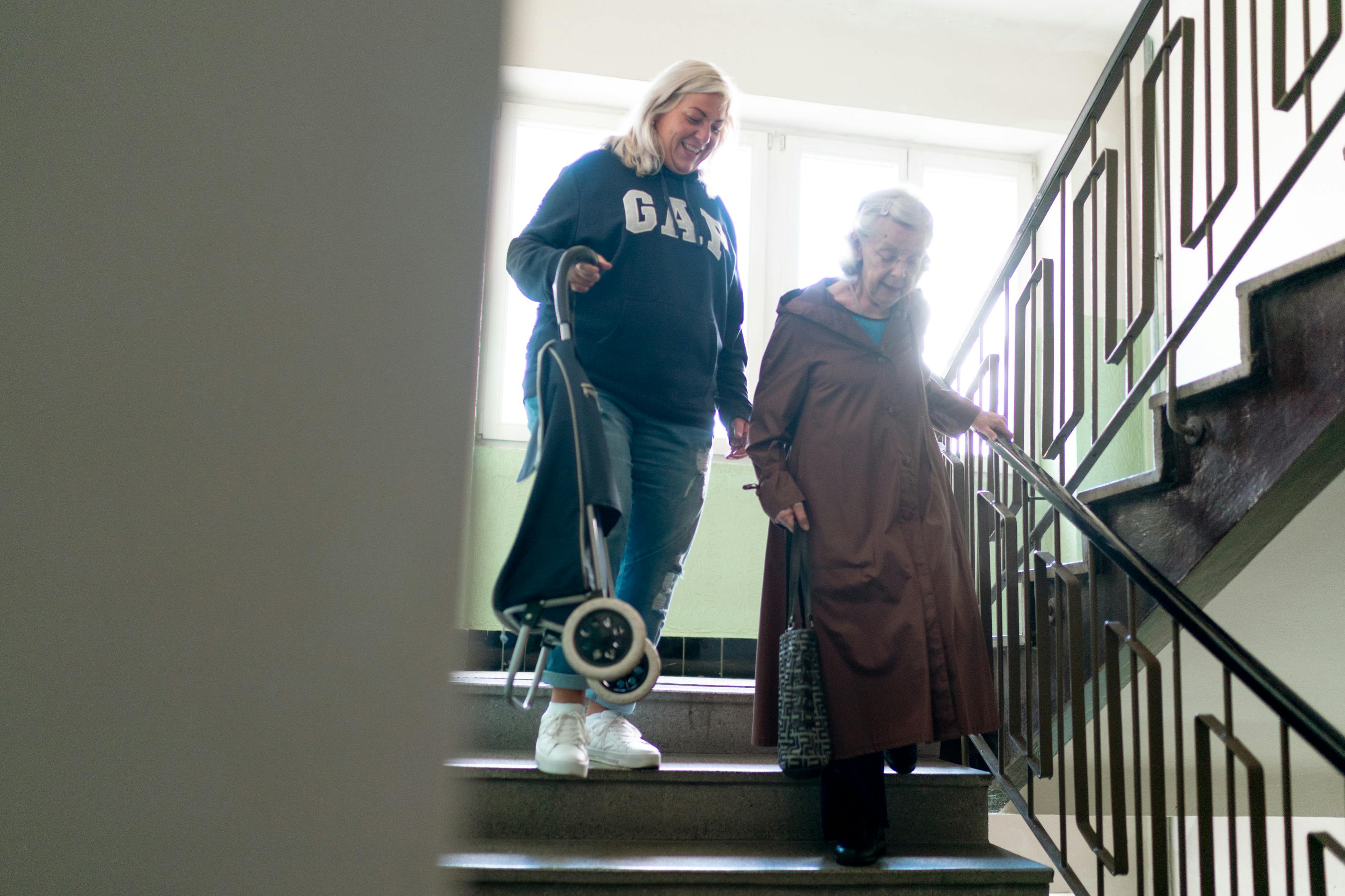 A caregiver helping an elderly woman down a stairwell with handrail support — the kind of fall-risk scenario that aging-in-place modifications (grab bars, ramps, stair lifts, lighting) are built to prevent in 2026.