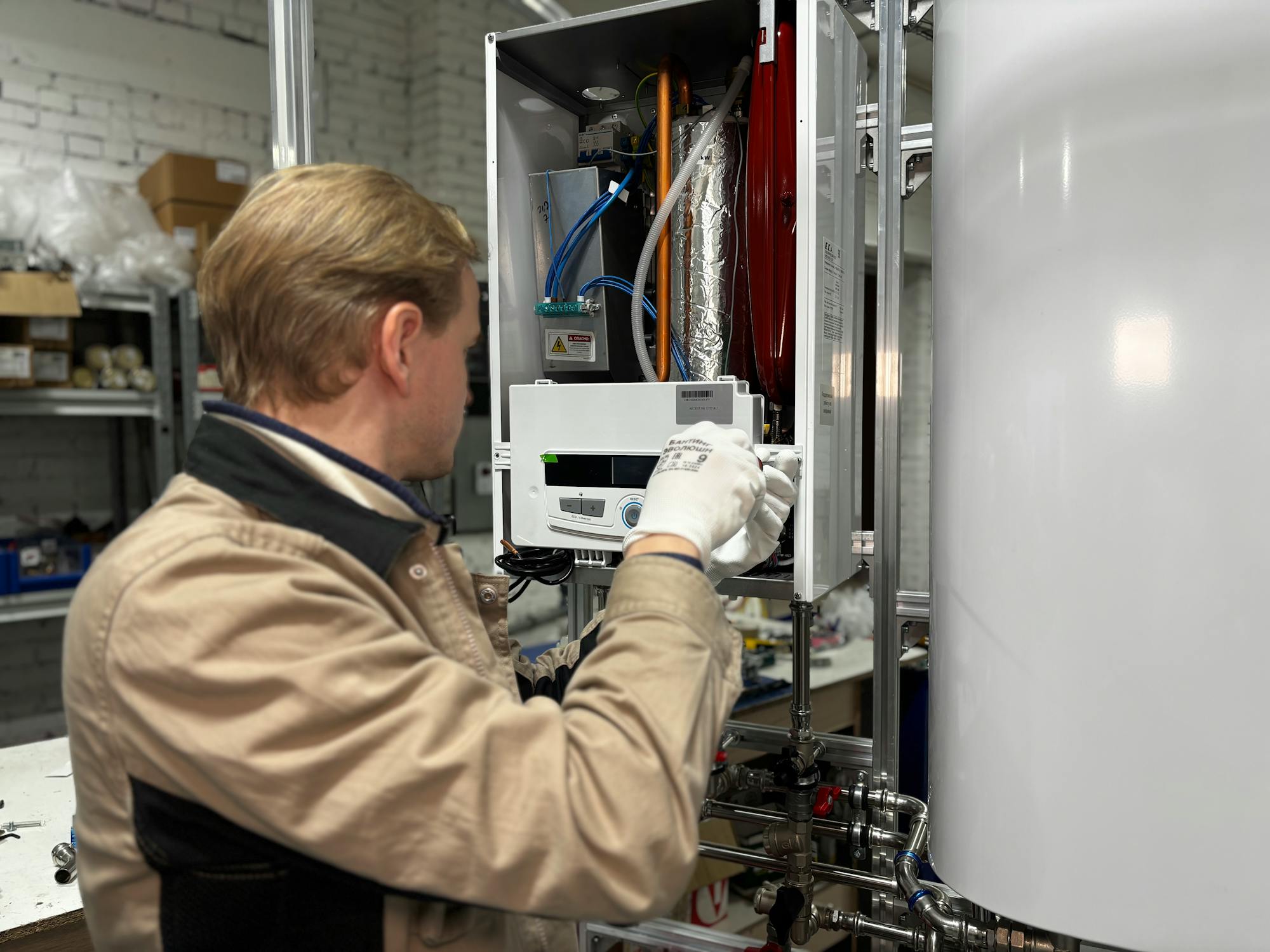 A plumber installing a modern residential water heater system — control panel commissioning with the white storage tank visible, copper supply lines and brass valves running below. The kind of $1,500-$8,000 high-margin install work that shifts a service-call shop's revenue mix toward replacements.