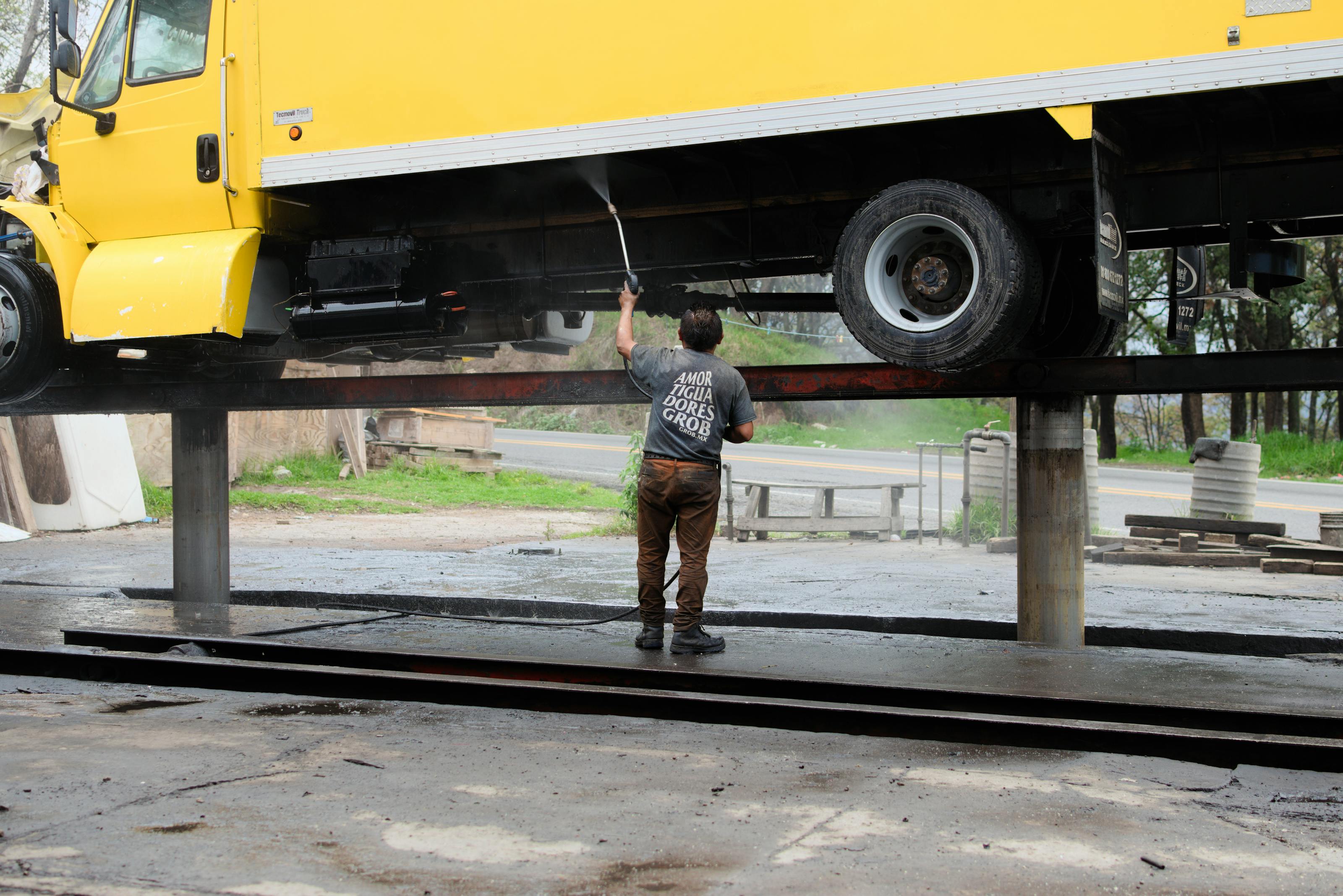 A pressure washing technician cleaning the underside of a raised yellow box truck at a fleet service yard — the kind of recurring commercial account that converts a residential pressure washing business into a route-density commercial operation in 2026.