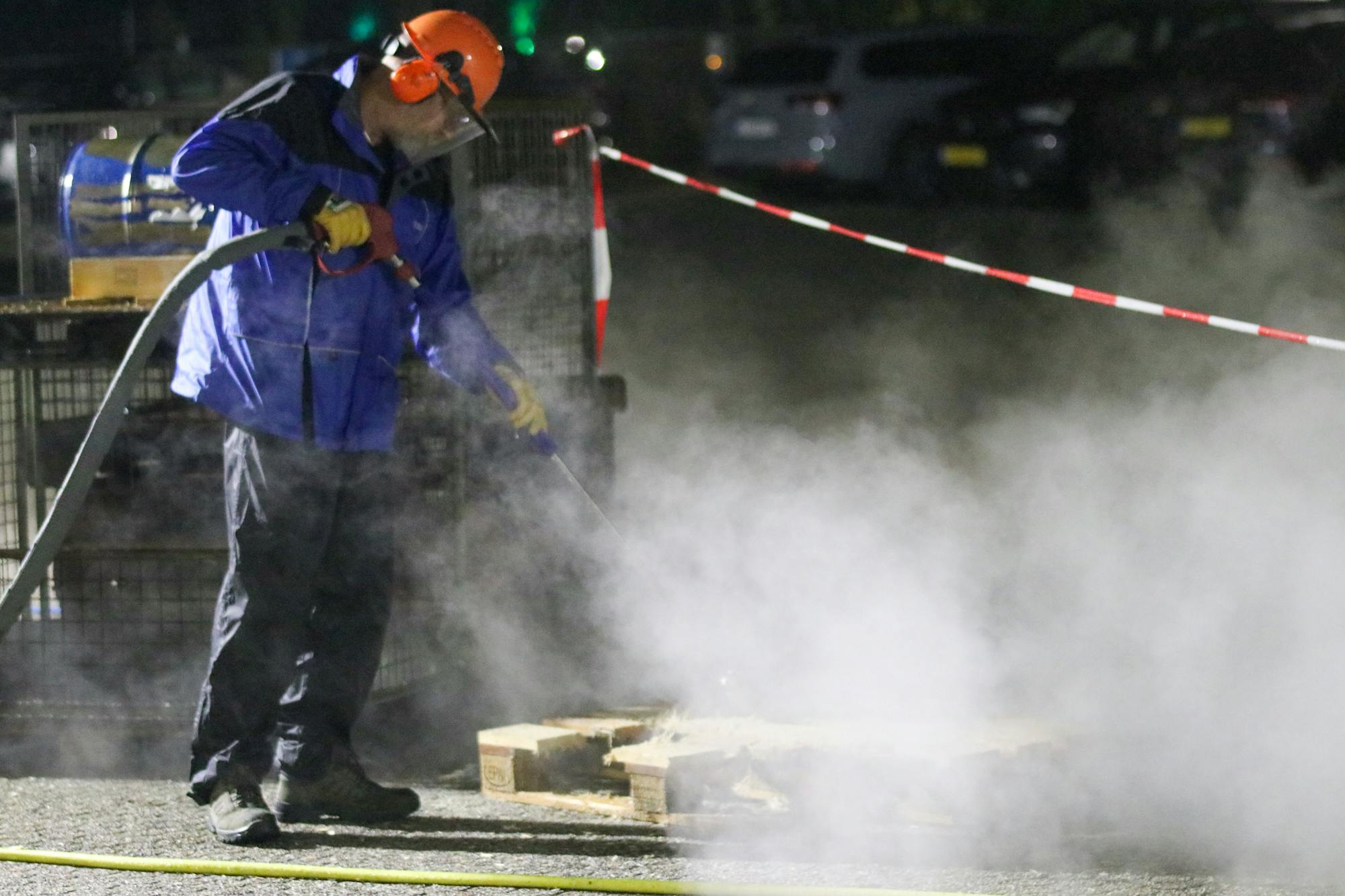 A pressure washing professional in full PPE (hard hat with face shield, chemical-resistant rain suit) working a high-pressure wand with steam visible — the kind of hot-water commercial cleaning work that runs 30-50% premium pricing over residential cold-water in 2026.