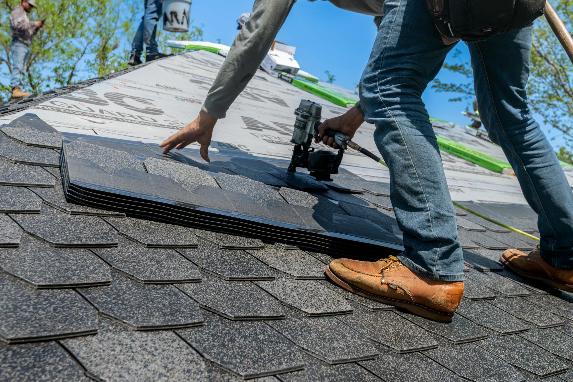 A roofer installing architectural asphalt shingles with a pneumatic coil nailer on a residential roof — the kind of $8,000-$15,400 average reroof work that anchors a residential roofing business in 2026.