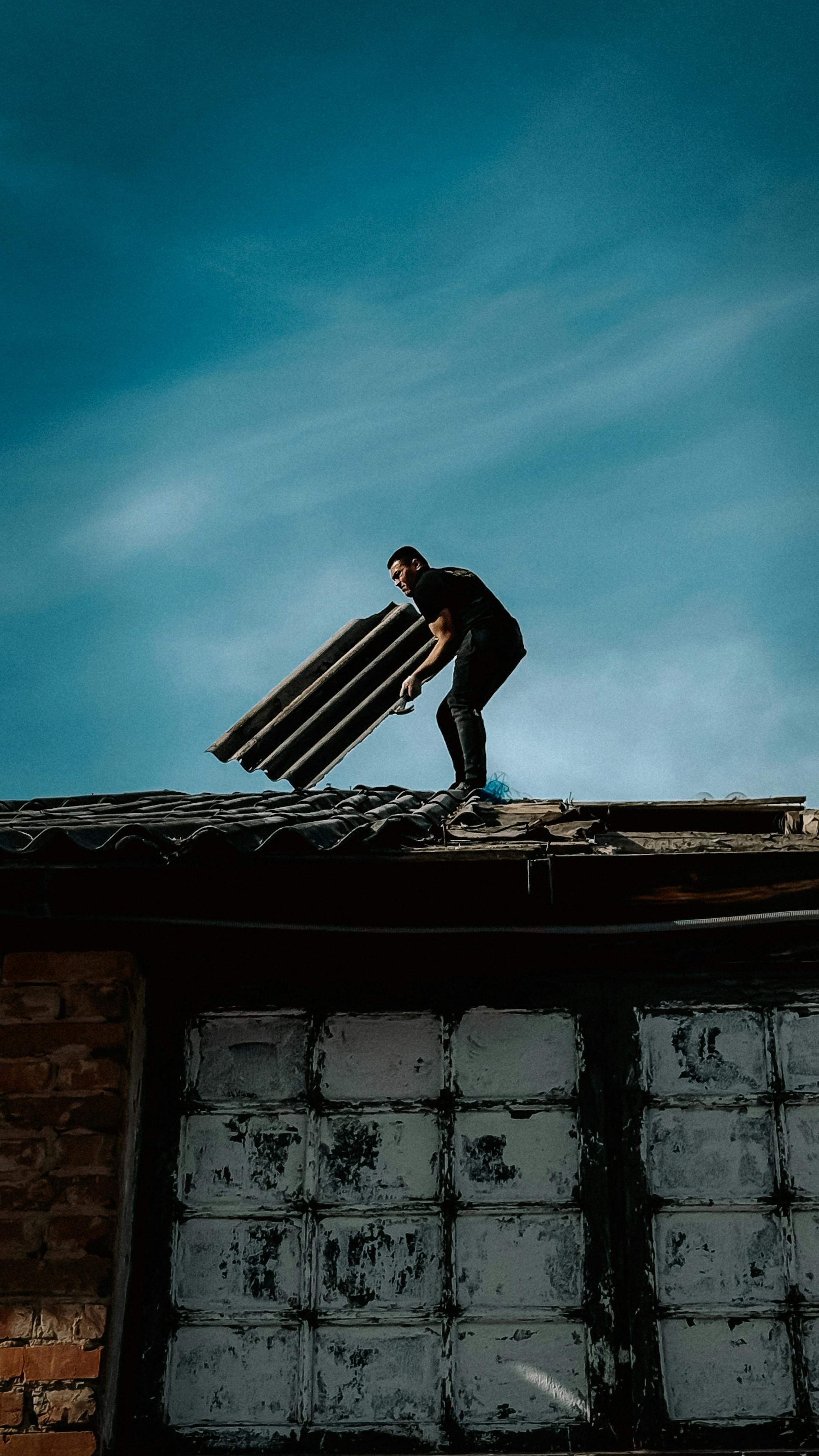 A roofer carrying metal sheeting on a residential roof under a stormy blue sky — the post-storm emergency tarp + restoration work that drives the dominant share of US residential roofing revenue in storm-prone markets in 2026.